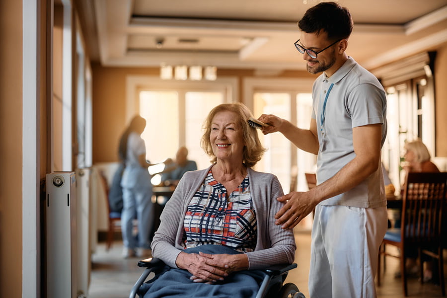 Happy senior woman in wheelchair with male nurse brushing her hair at residential care home.